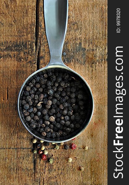 Black peppercorns in a metal measuring cup on a rustic wood table.