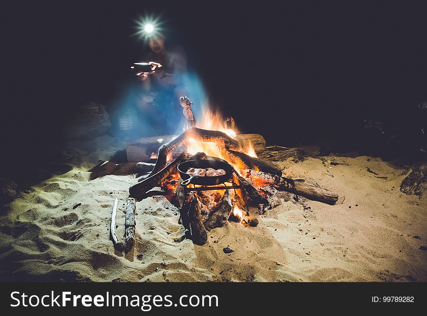 Person wearing headlamp sitting next to campfire on beach with skillet of cooking food. Person wearing headlamp sitting next to campfire on beach with skillet of cooking food.