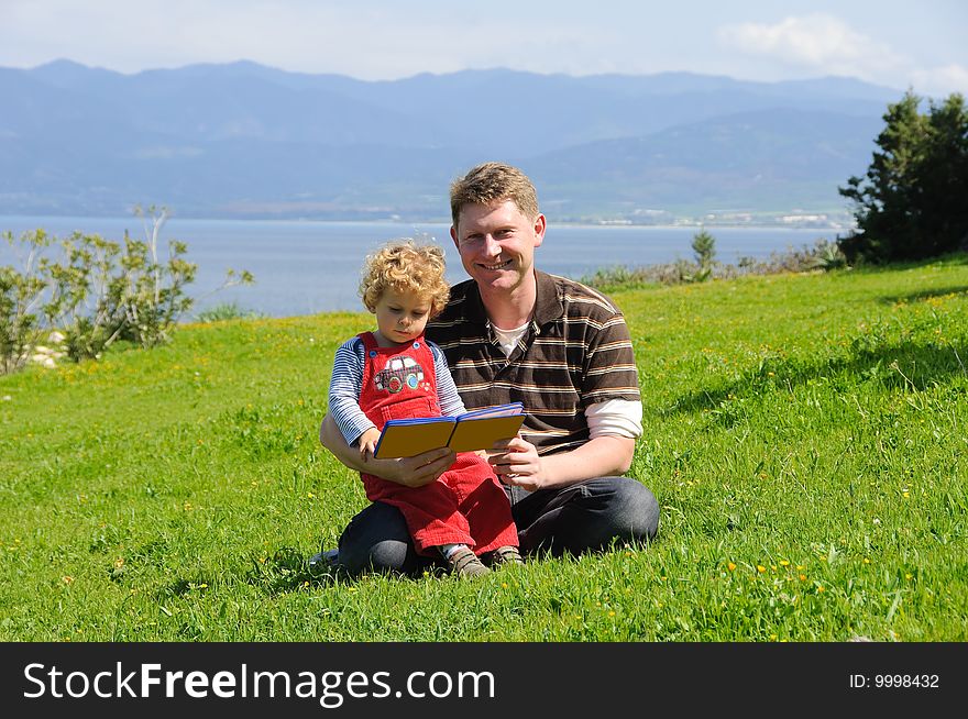 Father and son reading a book sitting on a green grass