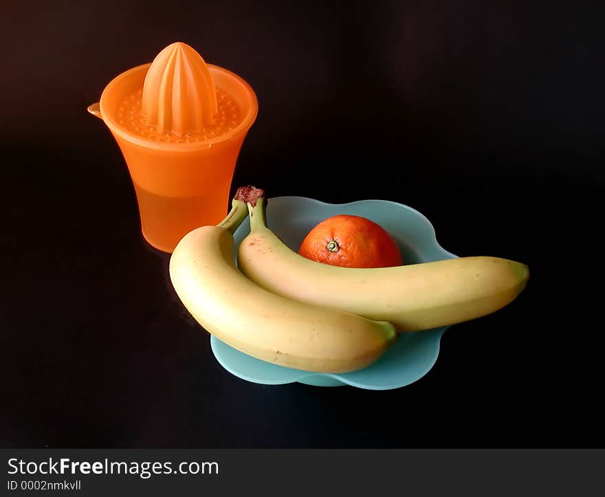 A still life composition with two fresh bananas on a blu dish and one orange, with a squeezer cup, on a black background. A still life composition with two fresh bananas on a blu dish and one orange, with a squeezer cup, on a black background