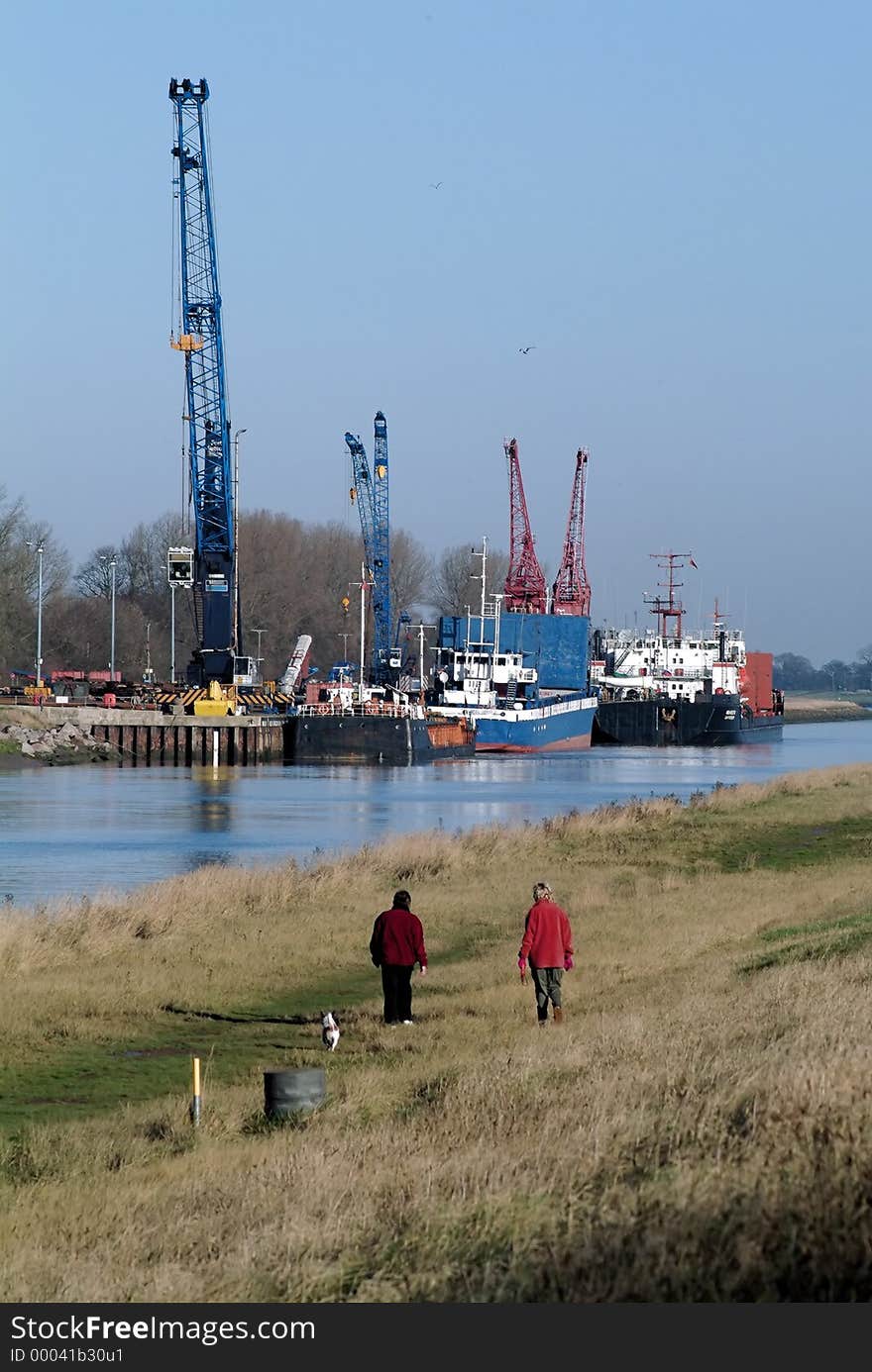 Two people walking a dog along a riverside path with a quay, ships, cranes in the background, winter, with clear blue sky backdrop. Taken at Sutton Bridge, Lincolnshire, England, UK. Two people walking a dog along a riverside path with a quay, ships, cranes in the background, winter, with clear blue sky backdrop. Taken at Sutton Bridge, Lincolnshire, England, UK