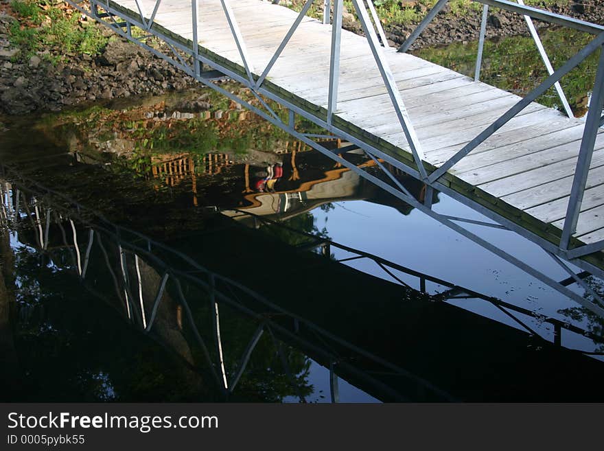 A picture of a foot bridge spanning the gap between the shore and a boat dock with the reflections showing in the dark water beneath. This was taken early one morning before the sun was overhead. A picture of a foot bridge spanning the gap between the shore and a boat dock with the reflections showing in the dark water beneath. This was taken early one morning before the sun was overhead.