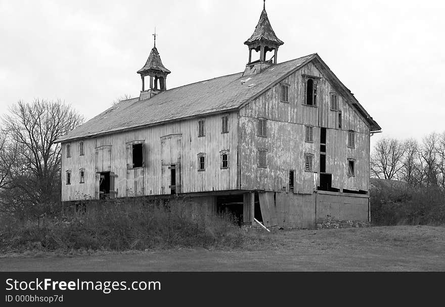 A large, very old barn in disrepair photographed in black and white. Many boards are missing and shingles fall from the roof as the barn slowly over the years decays. Of interest are the steeples on the roof, symbols of a successful farm in the days when this barn was first built. A large, very old barn in disrepair photographed in black and white. Many boards are missing and shingles fall from the roof as the barn slowly over the years decays. Of interest are the steeples on the roof, symbols of a successful farm in the days when this barn was first built