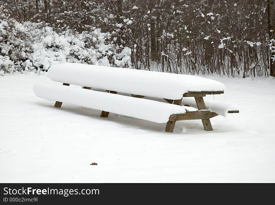 A long picnic table in a park covered by a heavy snowfall. A long picnic table in a park covered by a heavy snowfall.