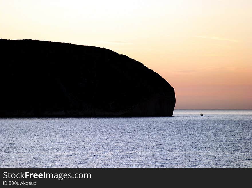 Small boat in the distance coming into a bay around a huge rock at sunrise in Spain. Small boat in the distance coming into a bay around a huge rock at sunrise in Spain