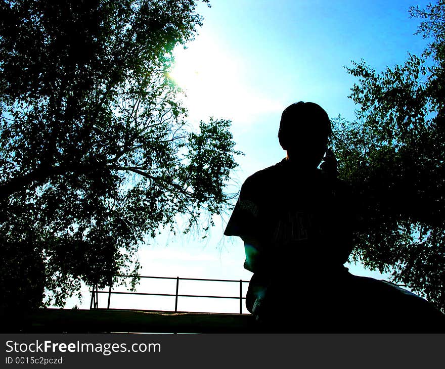 A teenage boy sits and is darkened by the sunlight. the background is of trees also blacked out except for the tips where the sun hits them, there they are green. the sky is a beautiful clear blue. A teenage boy sits and is darkened by the sunlight. the background is of trees also blacked out except for the tips where the sun hits them, there they are green. the sky is a beautiful clear blue.