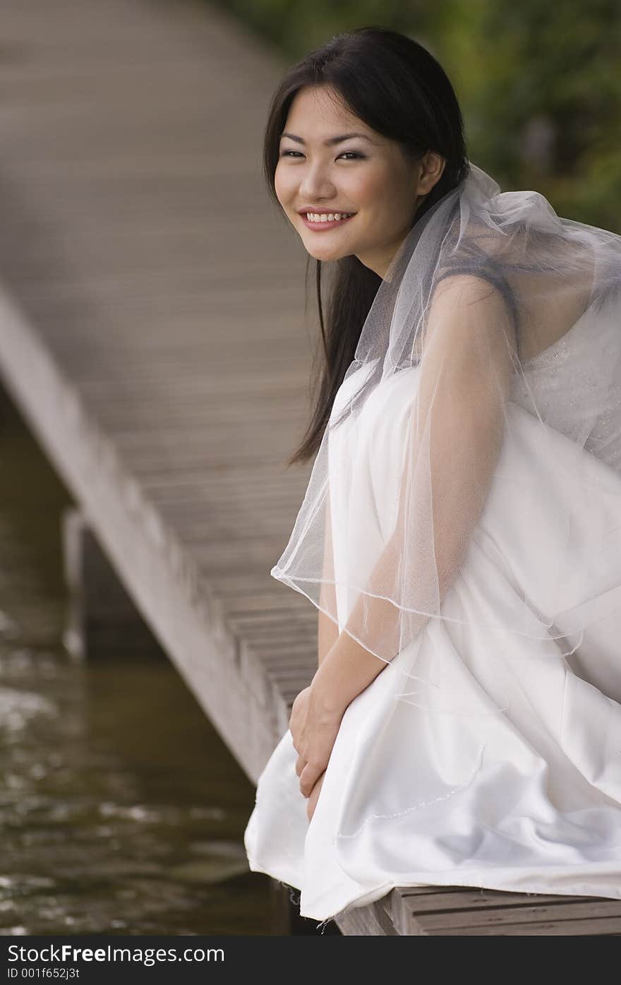 A beautiful asian bride sits on a boardwalk at the edge of a lake. A beautiful asian bride sits on a boardwalk at the edge of a lake