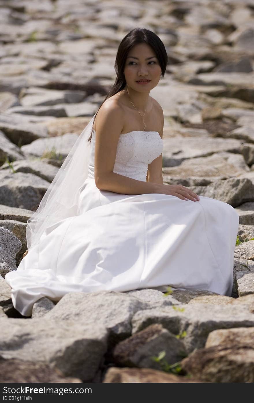 A beautiful asian woman sits on a rocky beach in a wedding dress. A beautiful asian woman sits on a rocky beach in a wedding dress