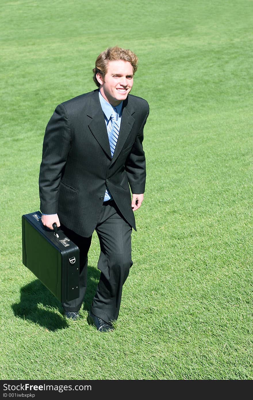 Young executive with briefcase in hand, chugs his way uphill with smile on all green grass. Young executive with briefcase in hand, chugs his way uphill with smile on all green grass