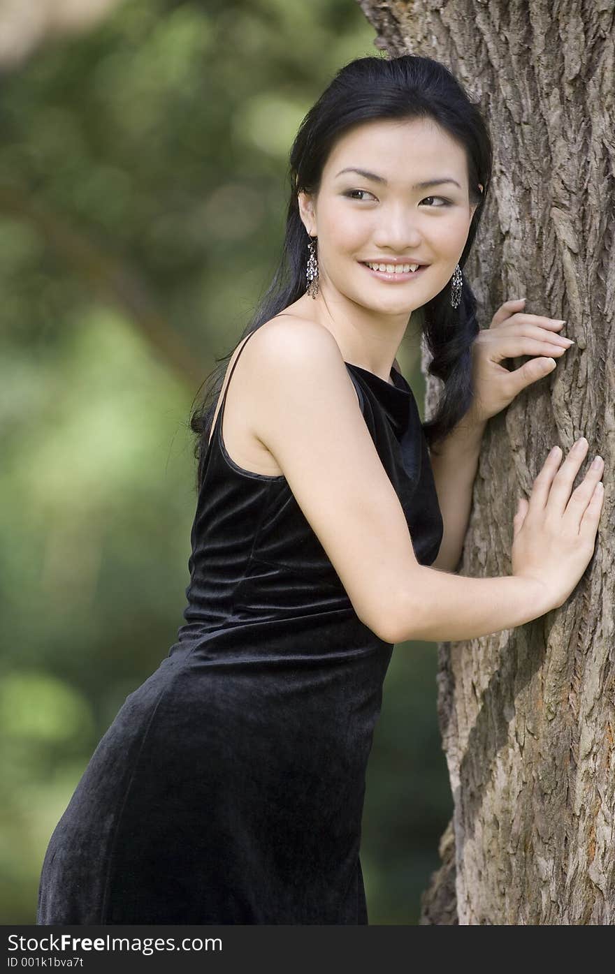 A beautiful asian model poses leaning against a tree in her black evening gown. A beautiful asian model poses leaning against a tree in her black evening gown