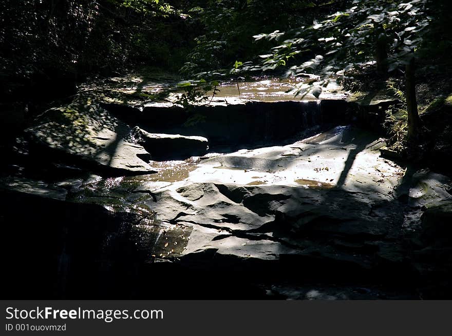 A splash of light falls on a very dark area of a stream, revealing a solid rock bed with a small stream running over it