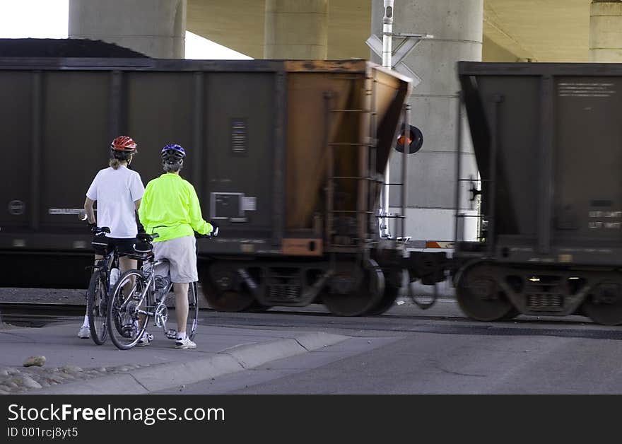 A pair of bikers wait on the train passing by. Train shows movement, bikers wait.