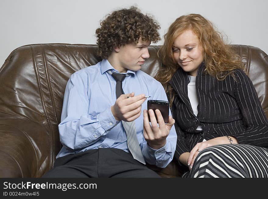 (Boy in relaxed office attire) Boy sitting on a leather couch is showing a girl beside him something on his palmtop. (Boy in relaxed office attire) Boy sitting on a leather couch is showing a girl beside him something on his palmtop