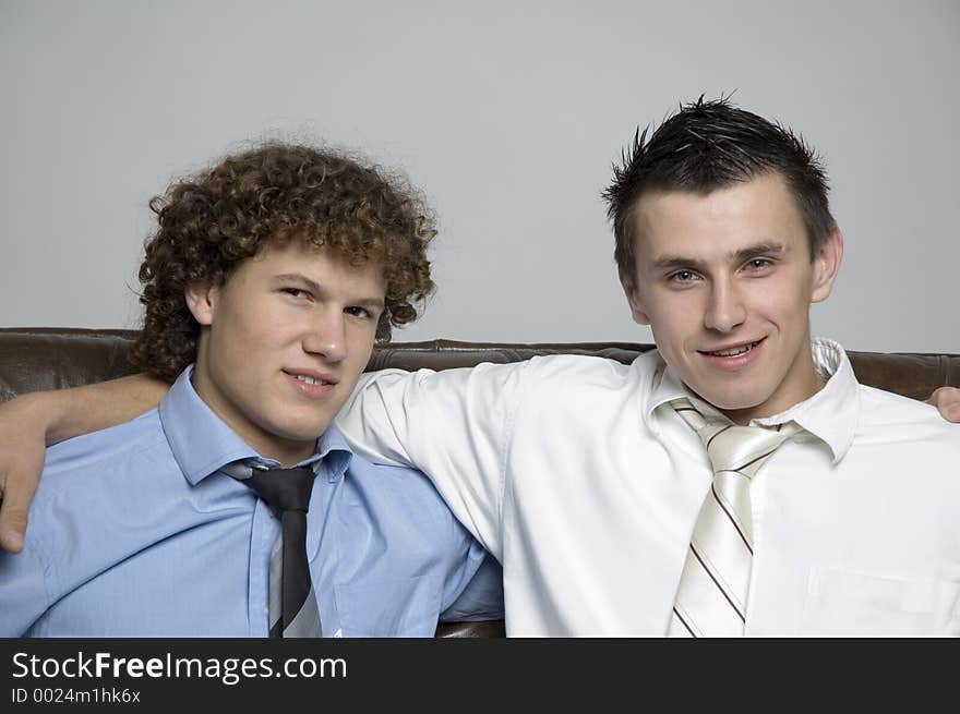 Two boys in relaxed business attire sitting on a couch together looking at the camera. Two boys in relaxed business attire sitting on a couch together looking at the camera