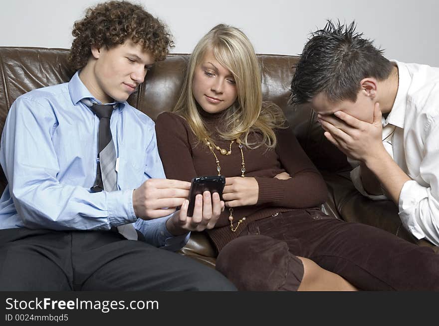 Two boys sitting beside a girl on a leather couch-one boy is showing her something on a palmtop while the other boy is burying his face in his hands. (Boys in relaxed office attire). Two boys sitting beside a girl on a leather couch-one boy is showing her something on a palmtop while the other boy is burying his face in his hands. (Boys in relaxed office attire)