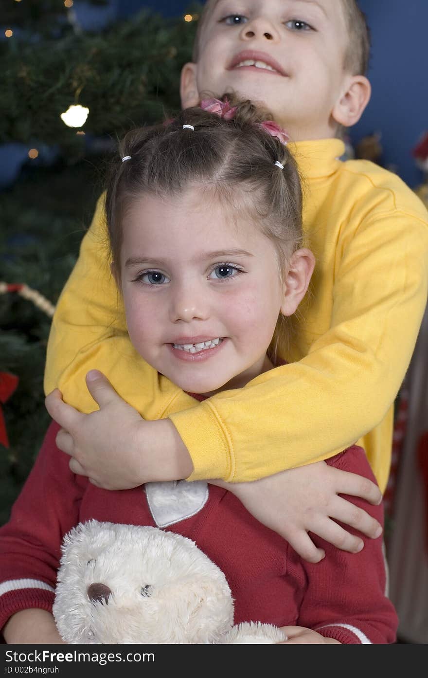 In front of a Christmas tree, boy stands behind his sister hugging her neck, the sister is holding a teddy bear. In front of a Christmas tree, boy stands behind his sister hugging her neck, the sister is holding a teddy bear