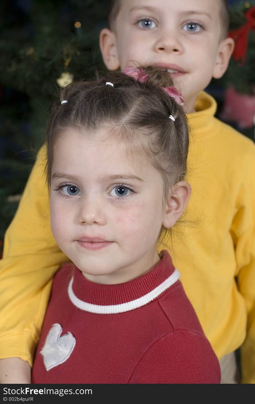 In front of a Christmas tree, boy stands behind his sister with his arm around her while she turns sideways and looks into the camera. In front of a Christmas tree, boy stands behind his sister with his arm around her while she turns sideways and looks into the camera