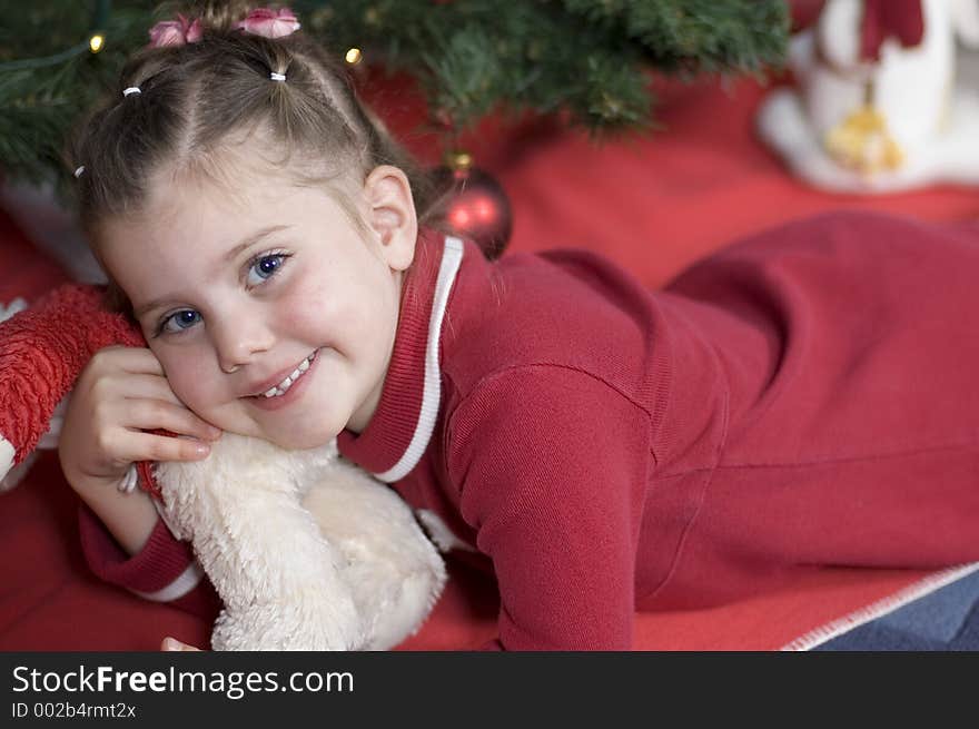 In front of a Christmas tree a little girl lies on her belly hugging her teddy bear. In front of a Christmas tree a little girl lies on her belly hugging her teddy bear