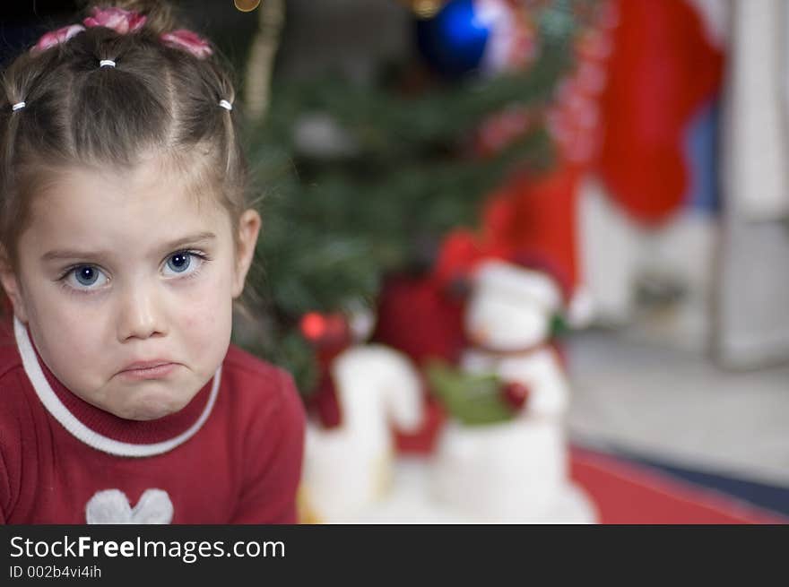 In front of a Christmas tree sits a girl who is frowning - Christmas snowmen in the background. In front of a Christmas tree sits a girl who is frowning - Christmas snowmen in the background