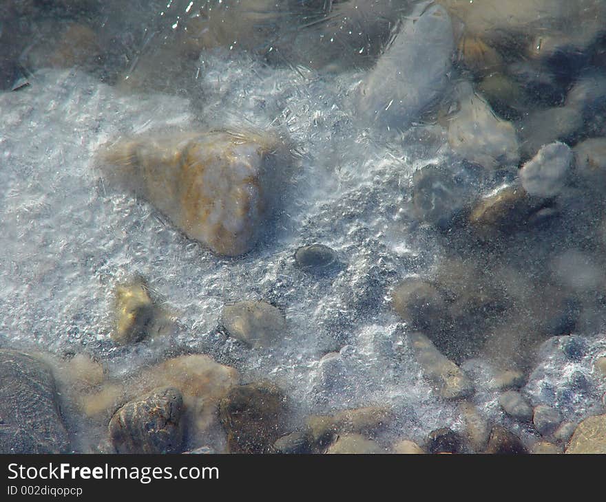 Shallows of a frozen river bed reveal ice, trapped air bubbles, cracks, patterns, multicolored rocks, great for background or texture. Shallows of a frozen river bed reveal ice, trapped air bubbles, cracks, patterns, multicolored rocks, great for background or texture