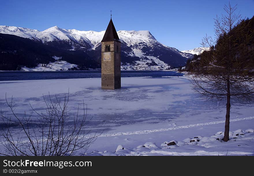 It shows a a bell tower of a church which is standing in a lake with view to Austria