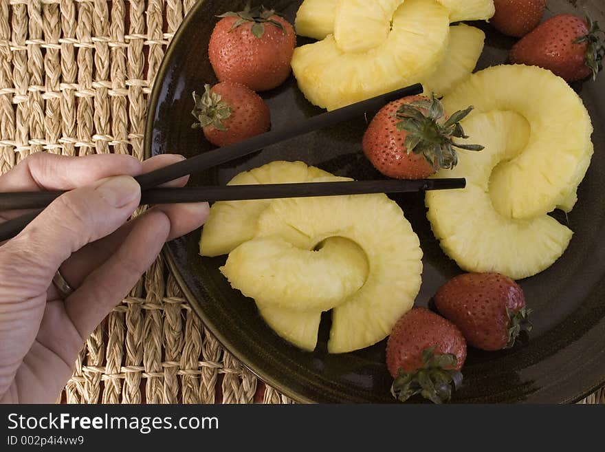 Close up of juicy yellow pineapple rings and red, plump strawberries and hand using chopsticks to pick up the strawberry. Close up of juicy yellow pineapple rings and red, plump strawberries and hand using chopsticks to pick up the strawberry.