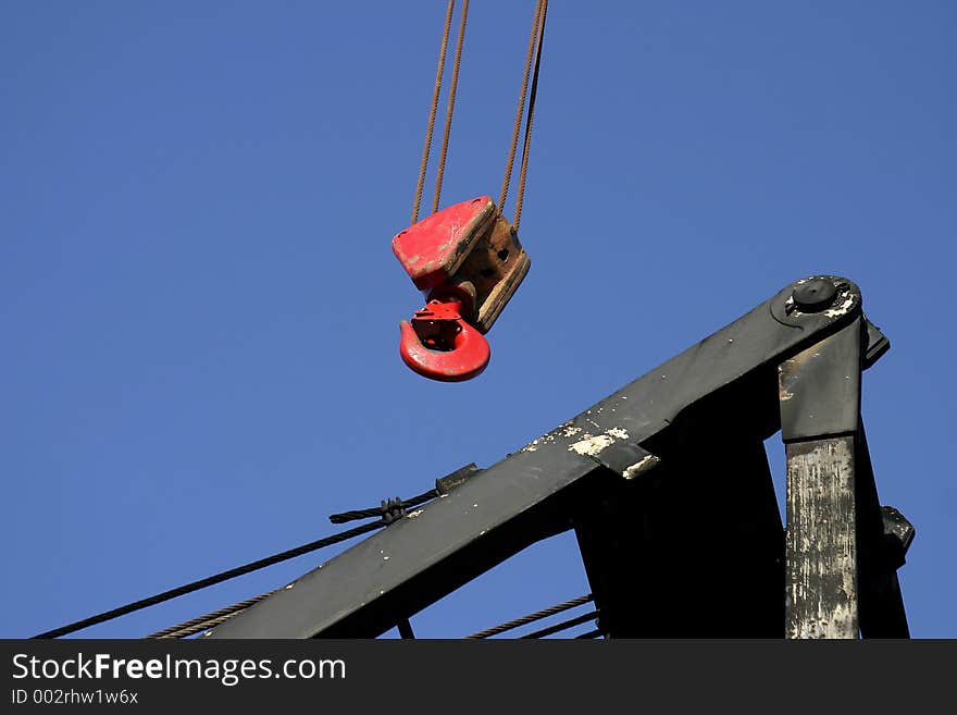 Red lifting hook suspended onfour twisted streel cables hanging over a black steel crane boom frame. Lots of blue sky for text. Red lifting hook suspended onfour twisted streel cables hanging over a black steel crane boom frame. Lots of blue sky for text.