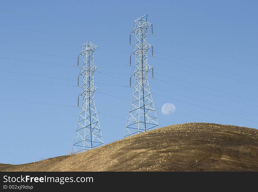 Hight voltage lines are held aloft by giant power towers, as the moon sets in the background. The hills appear scorched by a recent brush fire. Hight voltage lines are held aloft by giant power towers, as the moon sets in the background. The hills appear scorched by a recent brush fire.