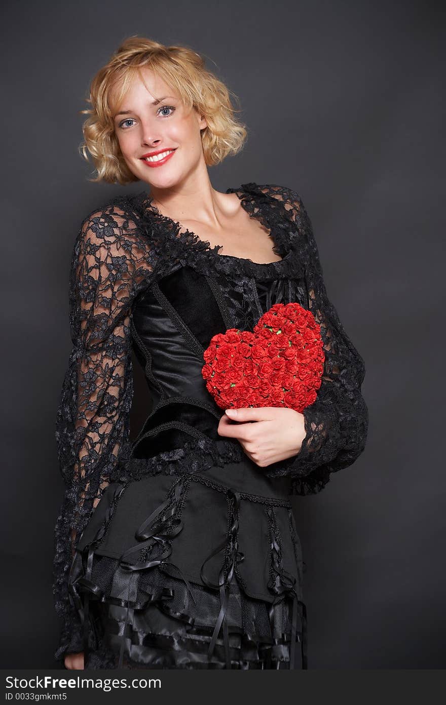 The beautiful blonde in an evening dress holds in hands heart made of roses. Background is black. The beautiful blonde in an evening dress holds in hands heart made of roses. Background is black