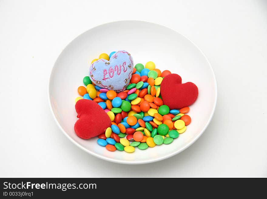 Three small cushioned hearts sprinkled on multi colored candied hearts in a white plate. Three small cushioned hearts sprinkled on multi colored candied hearts in a white plate