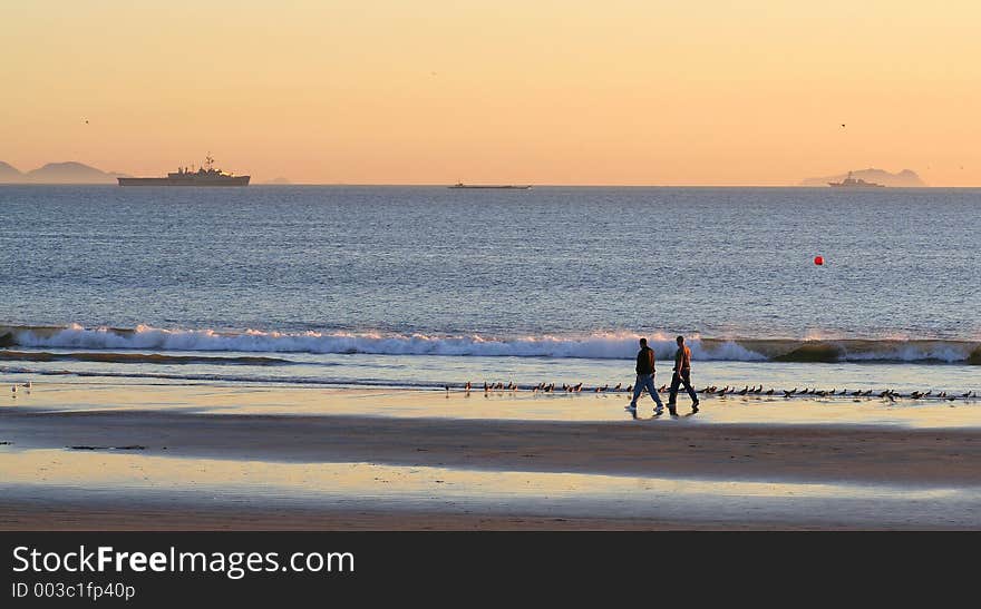 Two friends take a stroll on Southern California beach at sunset. Navy ships and Coronado Islands (Mexico) in the background. (Coronado, CA). Two friends take a stroll on Southern California beach at sunset. Navy ships and Coronado Islands (Mexico) in the background. (Coronado, CA)