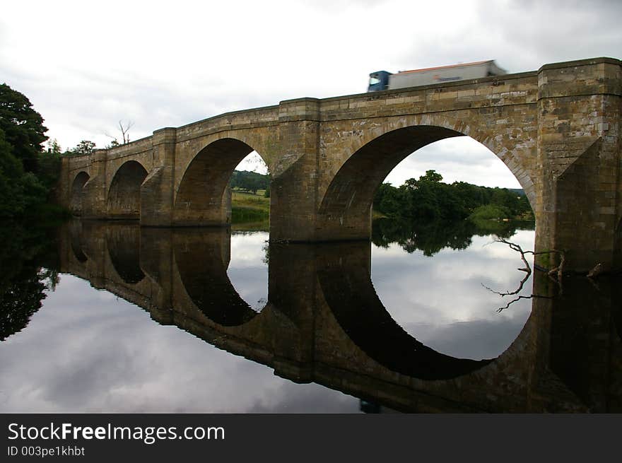 A truck driving over a bridge with a reflection in a river. A truck driving over a bridge with a reflection in a river