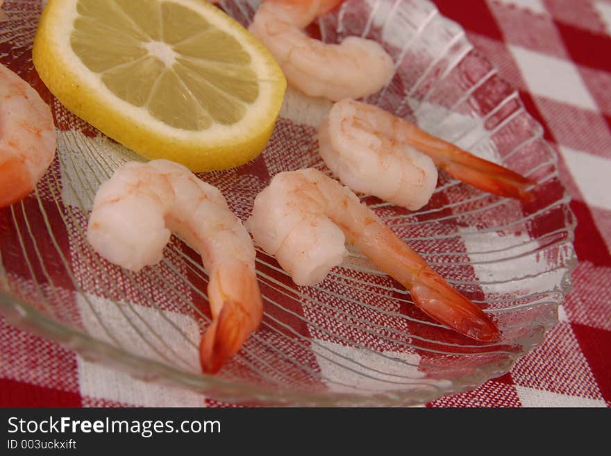 Boiled shrimp and lemon slice displayed on clear glass plate on red and white tablecloth.