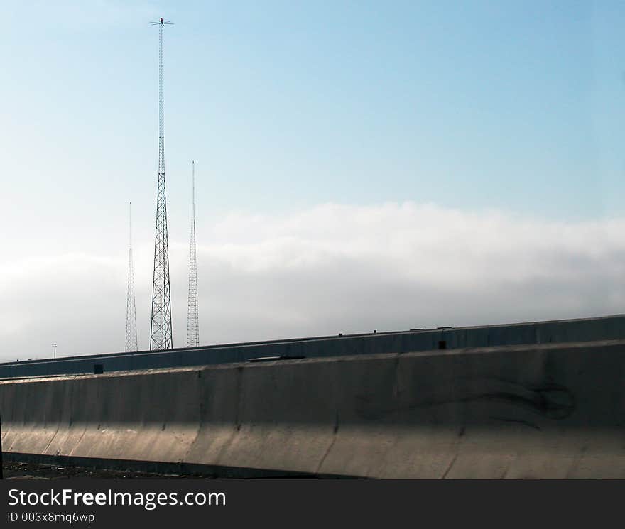 Viewed from a freeway tall radio towers are visible above concrete barriers and clouds. Viewed from a freeway tall radio towers are visible above concrete barriers and clouds