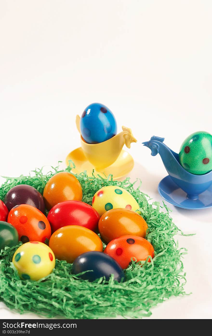 Coloured easter eggs in green grass with two eggcups looking like hens on white background