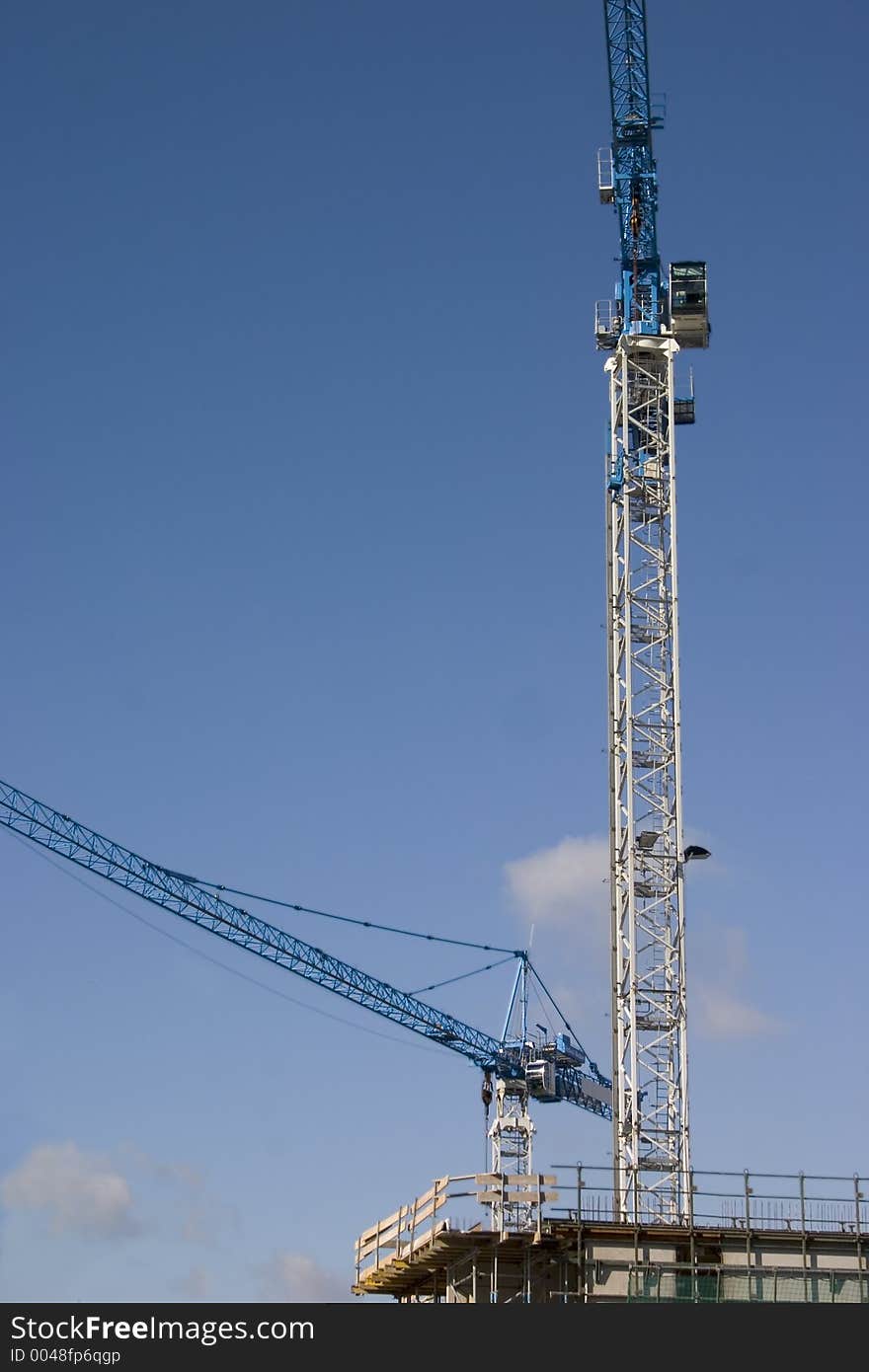 Two tall cranes tower over a construction site with blue sky in background. Two tall cranes tower over a construction site with blue sky in background