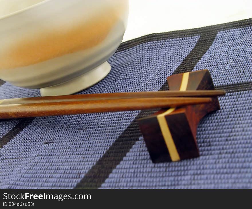 Perspective with chopsticks and bowl on a textile blue mat.Soft focus on the pointed end of the chopsticks,main focus in the middle of the image. Perspective with chopsticks and bowl on a textile blue mat.Soft focus on the pointed end of the chopsticks,main focus in the middle of the image.