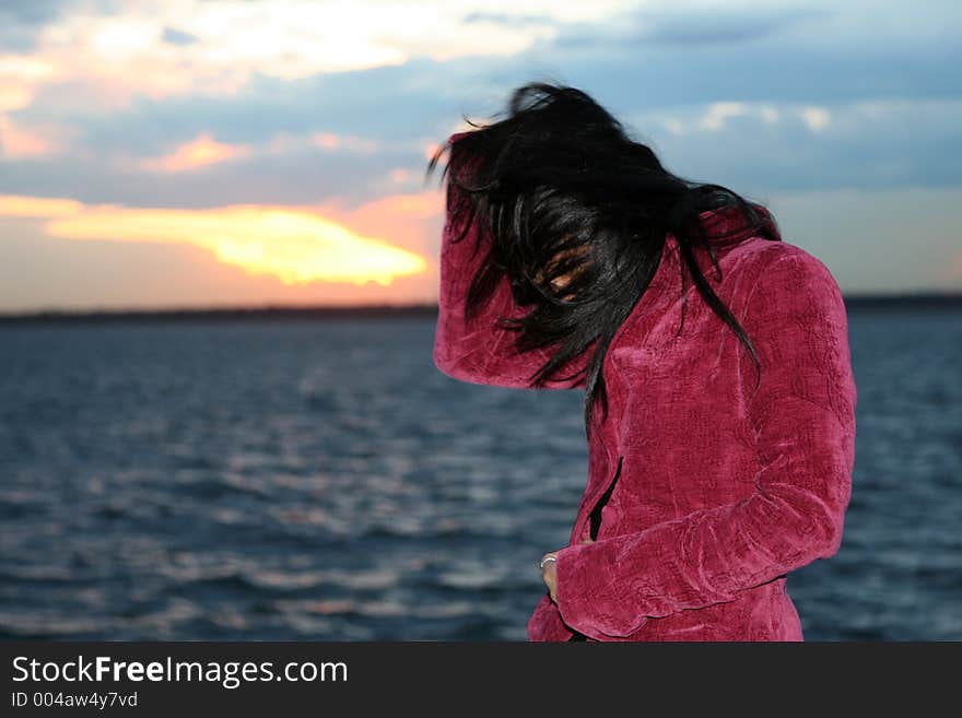 A girl in a jacket by the water at the sunset. Wind is blowing hair in her face. A girl in a jacket by the water at the sunset. Wind is blowing hair in her face.