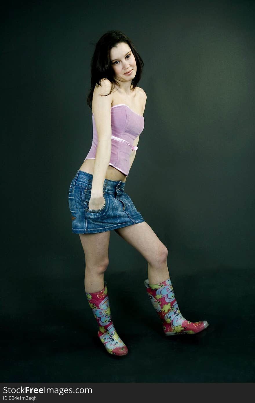 Young pretty brunette poses in the studio, dressed casual, with a light lilac top, denim skirt and colorful, funny plastic boots, trying various expressions, over a black background. Young pretty brunette poses in the studio, dressed casual, with a light lilac top, denim skirt and colorful, funny plastic boots, trying various expressions, over a black background