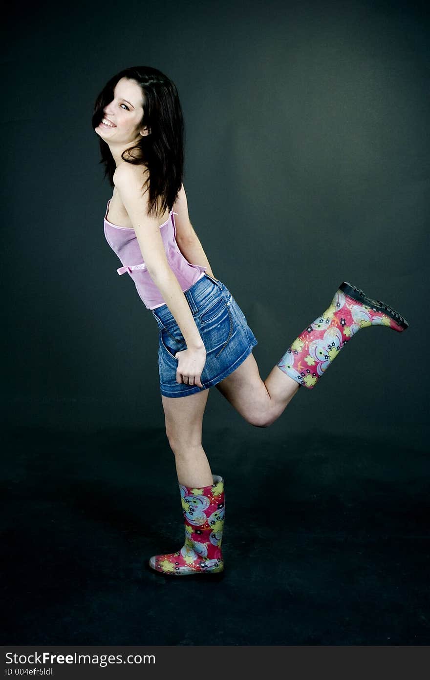 Young pretty brunette poses in the studio, dressed casual, with a light lilac top, denim skirt and colorful, funny plastic boots, trying various expressions, over a black background. Young pretty brunette poses in the studio, dressed casual, with a light lilac top, denim skirt and colorful, funny plastic boots, trying various expressions, over a black background