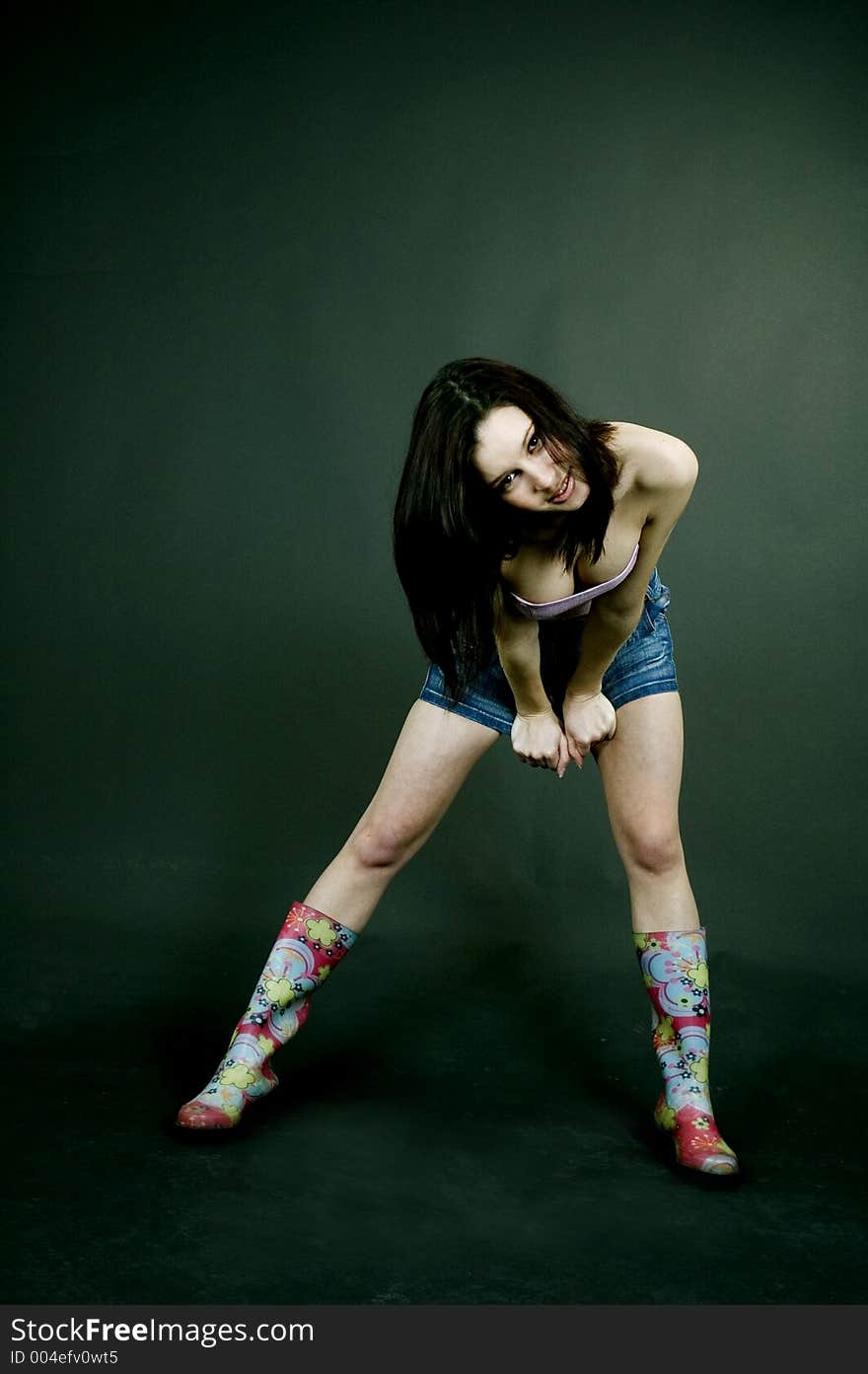 Young pretty brunette poses in the studio, dressed casual, with a light lilac top, denim skirt and colorful, funny plastic boots, trying various expressions, over a black background. Young pretty brunette poses in the studio, dressed casual, with a light lilac top, denim skirt and colorful, funny plastic boots, trying various expressions, over a black background