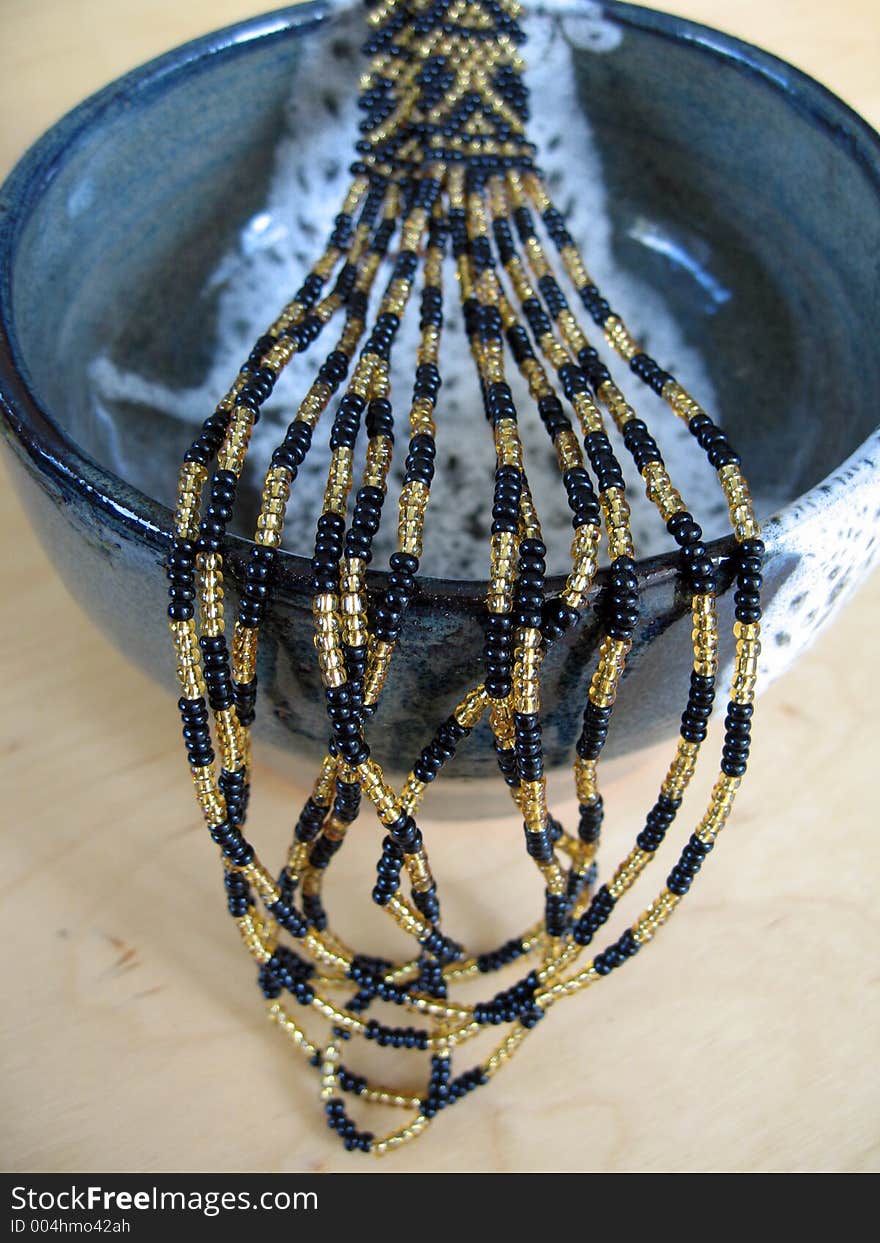 A black and gold beaded necklace presented over a ceramic bowl. This is an example of the craft of beading. The focus is on the beads close to the rim of the bowl. A black and gold beaded necklace presented over a ceramic bowl. This is an example of the craft of beading. The focus is on the beads close to the rim of the bowl.