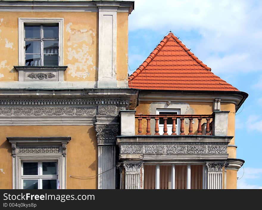 Fragment of a classic building, with pretty balcony with rail, and red roof. Peeling yellow paint and blue sky. Sculpted frieze. Fragment of a classic building, with pretty balcony with rail, and red roof. Peeling yellow paint and blue sky. Sculpted frieze.