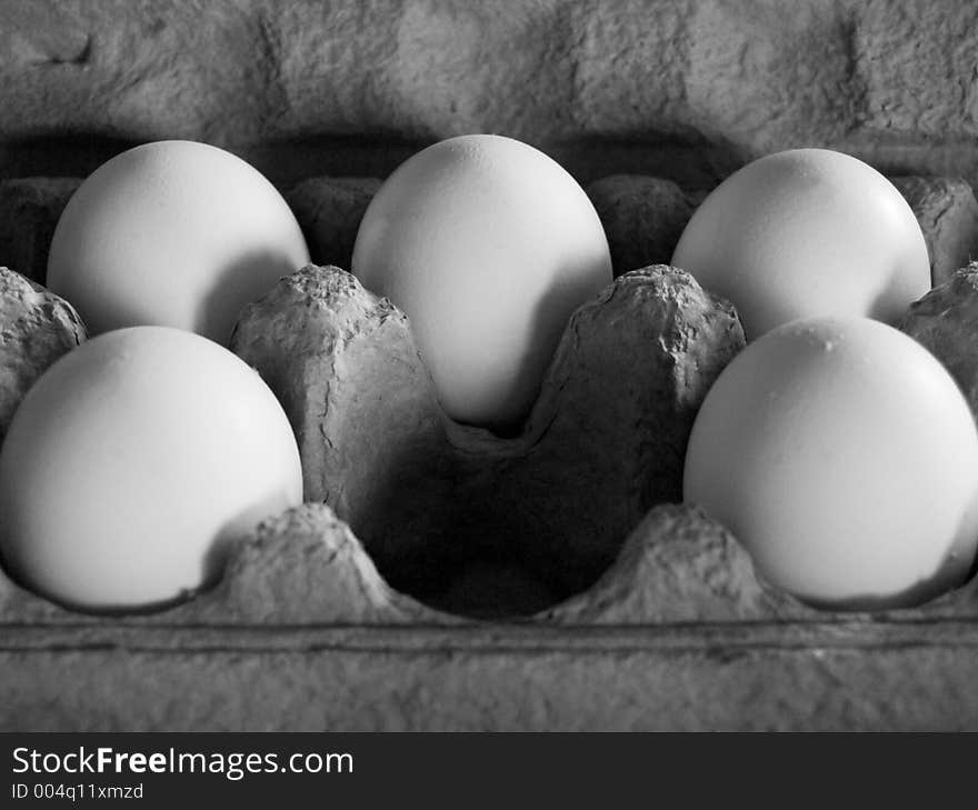 Five eggs in soft, dim light in a grey carton, arranged as to imply that a central egg has been removed. Grescale image.