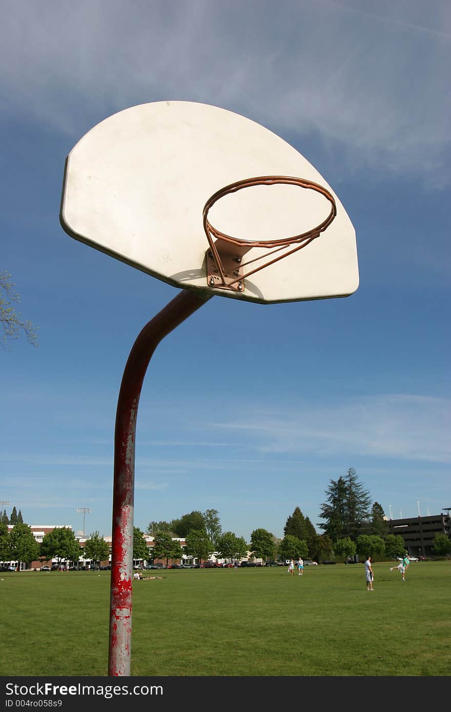 Rural basketball hoop without net and fields with playing kids in background.