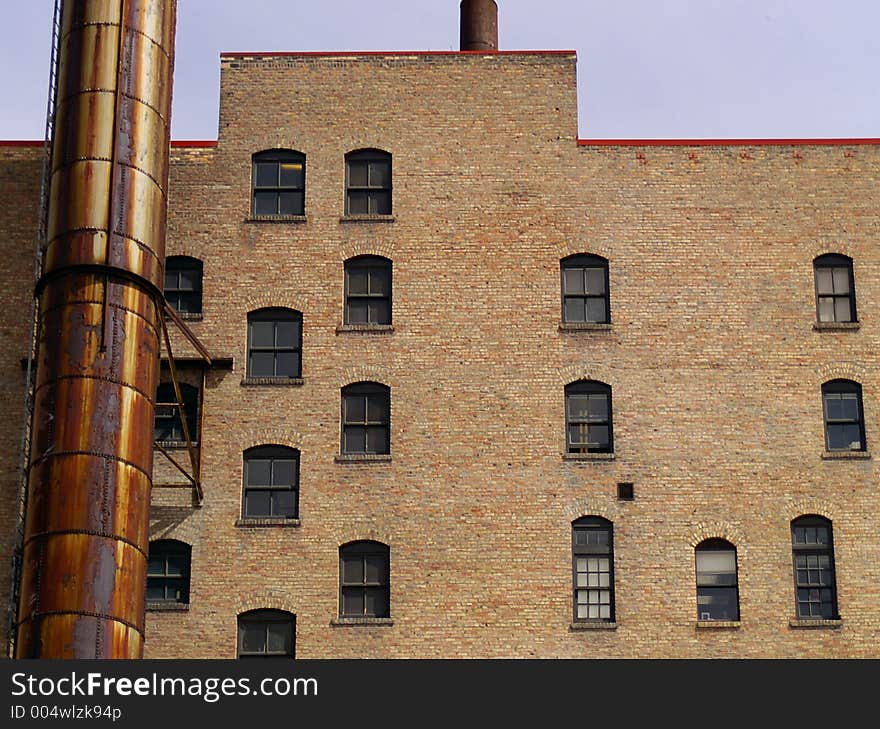 Windows and a rusted smokestack against a brown or tan brick wall on the side of an urban apartment building.