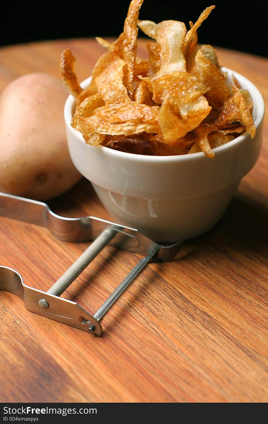 Crispy home-made french fries or potato chips served in a white ceramic bowl, on wooden chopping block with peeler and potato. Crispy home-made french fries or potato chips served in a white ceramic bowl, on wooden chopping block with peeler and potato.