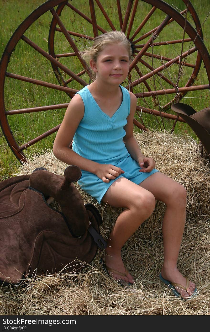 Girl sitting ontop of hay next to a saddle, wheel and hat. Girl sitting ontop of hay next to a saddle, wheel and hat