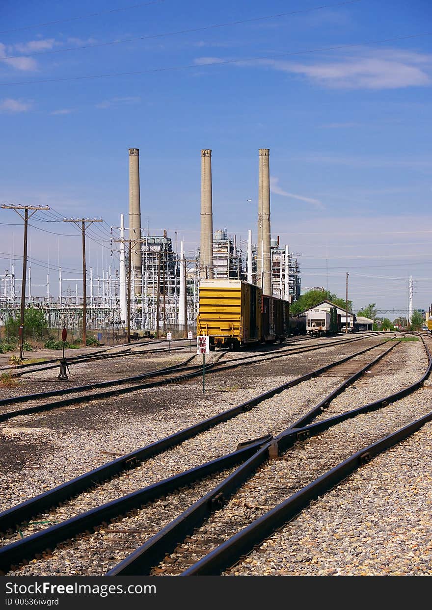 Rail lines run across a desolate industrial area with smokestacks and power lines in the background.