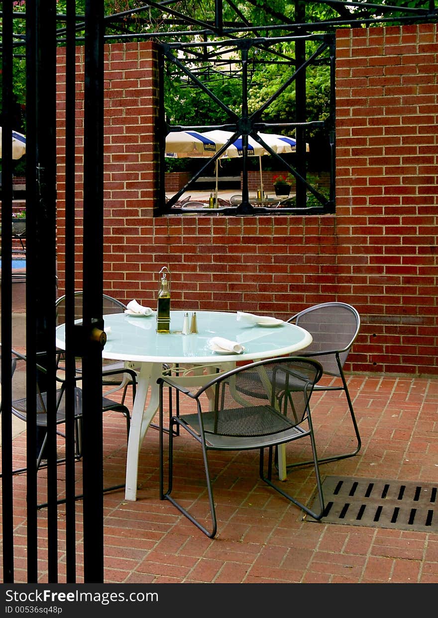 Secluded cyan dining table and chairs partially behind an iron gate at an outdoor cafe surrounded by red brick and green foliage.